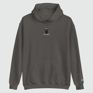 May include: A dark gray hoodie with a front pocket and a hood. The hoodie features a small graphic of a pint of stout with a shamrock and the text "VITAMIN G" printed below. The sleeve has a small embroidered logo.