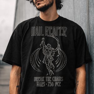May include: A black short-sleeved t-shirt featuring a white line-art graphic of a figure breaking chains while holding a scythe. The text reads HAIL REAPER, BREAK THE CHAINS, and MARS 736 PCE in a stylised font centred on the chest area.