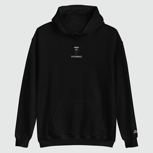 May include: Black hoodie with a front pocket and a hood. The hoodie features a small graphic of a drink with the text "VITAMIN G" printed below it. The sleeve has a small embroidered logo.