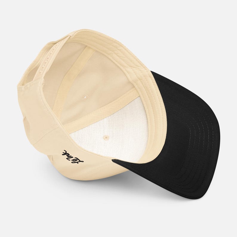 May include: A beige baseball cap with a black brim. The underside of the cap is white. The text "Le Ink" is written in black script on the side of the cap.