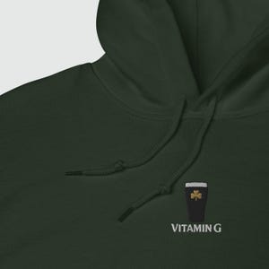 May include: Dark green hoodie with a drawstring hood. The hoodie features an embroidered design of a black pint glass with a gold shamrock and the text "VITAMIN G" in white.