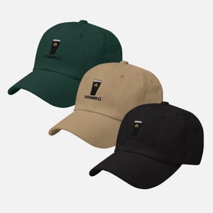 K&ouml;nnte beinhalten: Drei Baseballkappen in Dunkelgr&uuml;n, Beige und Schwarz. Jede Kappe hat eine schwarze Grafik eines Bierkrugs mit einem Kleeblatt und dem Text "VITAMIN G" auf der Vorderseite gestickt. Die Kappen haben einen gebogenen Schirm und einen verstellbaren Riemen.