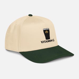 May include: A cream-colored baseball cap with a dark green brim and button. The cap features a black graphic of a pint of stout with a shamrock and the text "VITAMIN G" in black.
