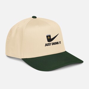 May include: A cream-colored baseball cap with a dark green brim and button. The cap features a black graphic of a beer mug with a shamrock and the words "JUST DRINK IT" in black.