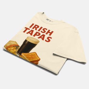 May include: Cream-colored t-shirt with the words "IRISH TAPAS" in red. The shirt features illustrations of a pint of dark beer, a bag of chips, and a stack of toasted bread. The design is a playful take on Irish cuisine.