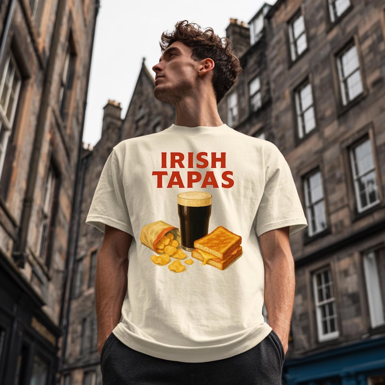 May include: Cream-colored t-shirt with the text "IRISH TAPAS" in red. The shirt features an illustration of a pint of stout, chips, and a grilled cheese sandwich. The model is standing in front of a stone building.