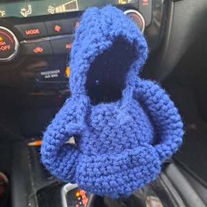 May include: A handmade, blue crochet hoodie car accessory. The miniature hoodie has a hood and sleeves, and is designed to fit over a car's gear stick. The crochet work is visible, with a textured appearance.