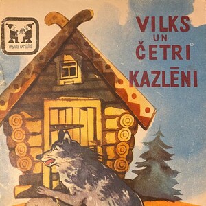 May include: A vintage children's book cover featuring a wolf in a blue-grey color, with a brown wooden house in the background. The title of the book is "VILKS UN ČETRI KAZLĒNI" in red lettering.