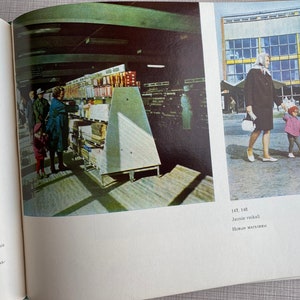 May include: A black and white photo of a grocery store interior with people shopping. The photo is from the 1960s or 1970s. The store is well-stocked with shelves full of products. The photo is taken from the perspective of someone standing in the aisle. The text in the photo reads "Jaunie veikali" and "Новые магазавы".