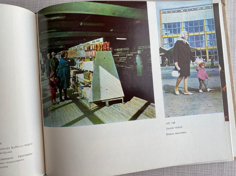 May include: A black and white photo of a grocery store interior with people shopping. The photo is from the 1960s or 1970s. The store is well-stocked with shelves full of products. The photo is taken from the perspective of someone standing in the aisle. The text in the photo reads "Jaunie veikali" and "Новые магазавы".