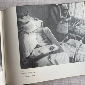 May include: A black and white photo of a nurse in a white uniform and hat holding a baby in a crib. The nurse is looking at the baby. There are other cribs in the background with babies in them. The text on the page reads "106 Mēs esam dzimu&scaron;i Rīgā Mēs radāmies Rīgā"