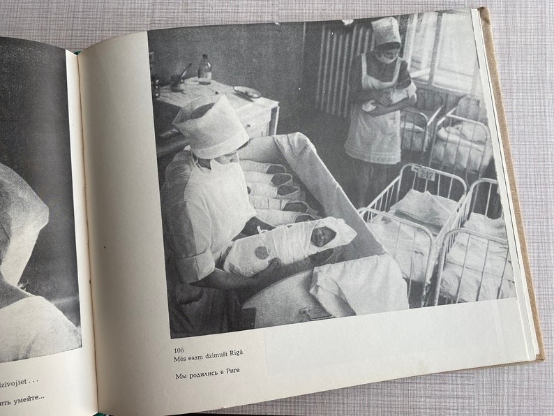 May include: A black and white photo of a nurse in a white uniform and hat holding a baby in a crib. The nurse is looking at the baby. There are other cribs in the background with babies in them. The text on the page reads "106 Mēs esam dzimu&scaron;i Rīgā Mēs radāmies Rīgā"