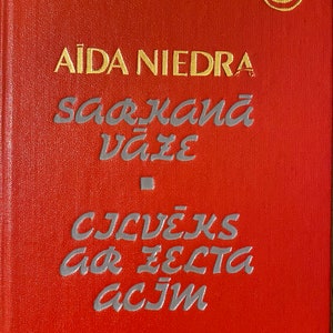 May include: A red hardcover book with gold and silver lettering. The title "Vakara Romāns" is at the top, with the author's name and the title "Sarkana Vāze" in the centre. The book has a gold emblem in the upper right corner.