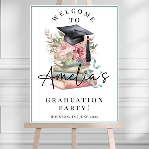 May include: A graduation party welcome sign on an easel. The sign features the text "Welcome to Amelia's Graduation Party!" with a black graduation cap, flowers, and books. The sign is in a white frame with a teal border.