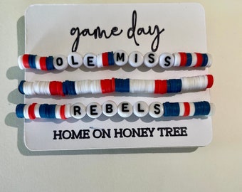 May include: Three white beaded bracelets with red, white, and blue accents. The bracelets spell out "OLE MISS" and "REBEL" with the text "game day" and "HOME ON HONEY TREE" on a white card.