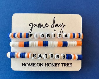 May include: Two white beaded bracelets with the words "FLORIDA" and "GATORS" on them. The bracelets are blue, orange, and white. The card behind the bracelets says "game day" and "HOME ON HONEY TREE".