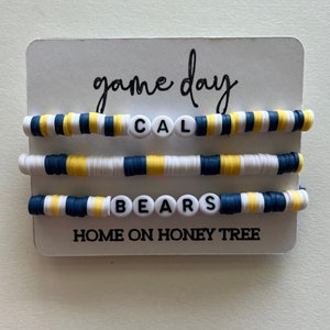 May include: Three white beaded bracelets with blue and yellow stripes. The bracelets spell out "CAL BEARS" and "HOME ON HONEY TREE".