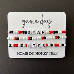 May include: Three beaded bracelets with the words "UTAH" and "UTES" in black letters on white beads. The bracelets are red, white, and black. The card reads "game day" and "HOME ON HONEY TREE".