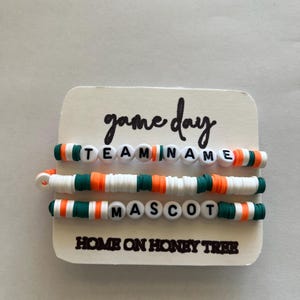 May include: Three beaded bracelets with white, orange, and green accents are displayed on a white card. The top bracelet spells out "TEAM NAME" with white letter beads. The card reads "game day" and "HOME ON HONEY TREE".