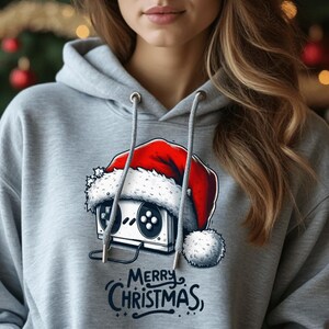 Può includere: Una felpa grigia con un'illustrazione bianca e rossa di una console per videogiochi retrò che indossa un cappello da Babbo Natale. Il testo "Merry Christmas" è stampato sotto l'illustrazione.