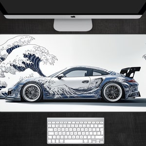 Puede incluir: Una alfombrilla de escritorio blanca con una ilustración azul y blanca de un coche y olas. El coche es un Porsche 911 GT3 RS con una pintura personalizada que presenta un diseño de olas. Las olas están en el estilo de una impresión japonesa tradicional en madera.