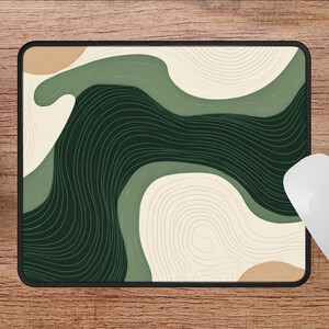 May include: Rectangular mouse mat with a black border, featuring an abstract design in shades of green, beige, and dark green. The design includes curved lines and shapes. A white mouse is next to the mat. The mat measures 22.86cm x 17.78cm.