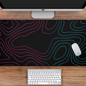 Pode incluir: Um mouse pad retangular preto com um design topográfico em tons de rosa e verde-azulado. O mouse pad tem 78,7 cm de comprimento e 39,4 cm de largura. Um mouse e um teclado de computador brancos também são visíveis.
