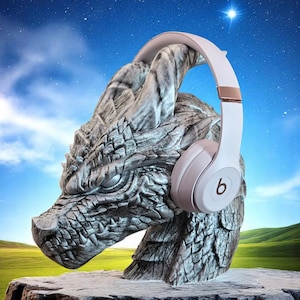Puede incluir: Unos auriculares blancos con un acento de oro rosa metálico en la diadema, con el logotipo de Beats en el auricular, colocados sobre una escultura de piedra gris de una cabeza de dragón con escamas y cuernos detallados.