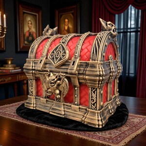 May include: A decorative chest with a red quilted interior and ornate gold and bronze accents. The chest features gargoyle and geometric designs, with a hinged lid and a central lock. The chest is placed on a patterned rug.