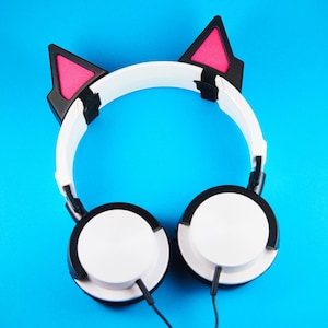 May include: White headphones with black accents and cat ear attachments, featuring pink interiors. The headphones have a white headband and earcups, with black cables. The background is a solid blue.
