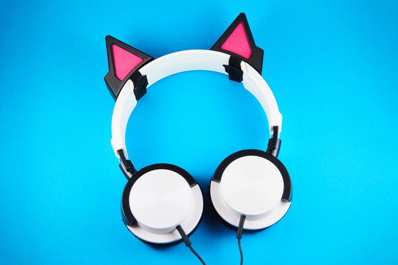 May include: White headphones with black accents and cat ear attachments, featuring pink interiors. The headphones have a white headband and earcups, with black cables. The background is a solid blue.