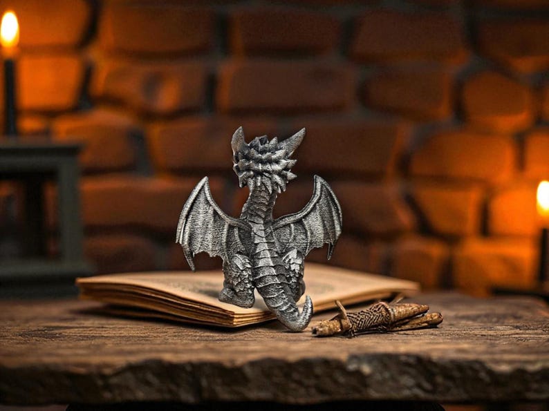 Puede incluir: Una detallada figura de drag&oacute;n plateado con alas extendidas, posada sobre un libro abierto. El drag&oacute;n est&aacute; intrincadamente esculpido, mostrando escamas y alas texturizadas. La escena est&aacute; ambientada sobre una superficie de madera con un fondo de ladrillo y velas encendidas.
