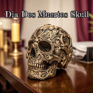 May include: An ornate gold and black skull sculpture with floral detailing, displayed on a wooden surface. The skull features intricate carvings and is labelled "Dia Dos Muertos Skull". The background includes candles and a dark red curtain.