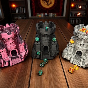Puede incluir: Tres torres de dados en miniatura con forma de castillo en rosa, negro y gris, cada una con un conjunto de dados diferente. Las torres tienen una textura similar a la piedra y están sobre una superficie de madera. Los dados son de varios colores.