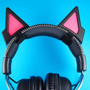 May include: Black headphones with cat ear attachments. The cat ears are black with pink inner ear details. The headband is black with a silver zipper detail. The background is a solid blue.