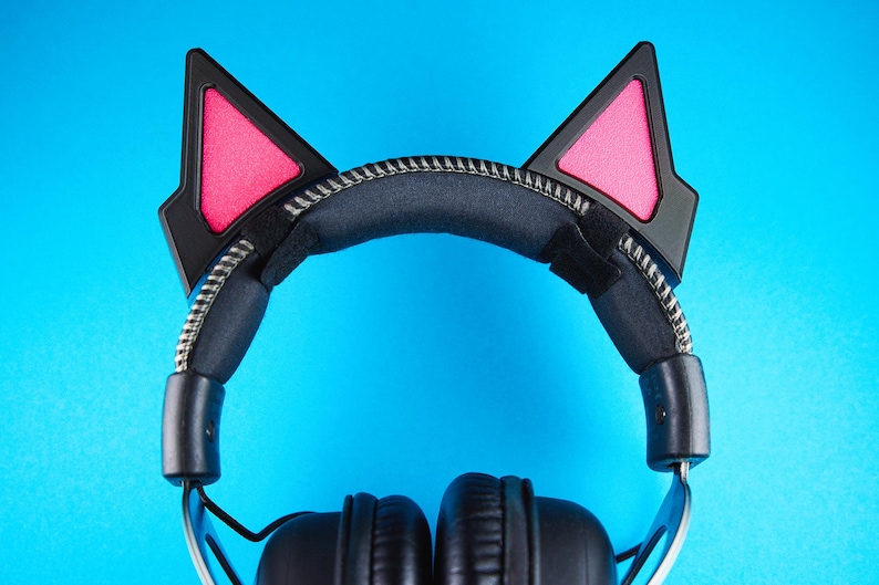 May include: Black headphones with cat ear attachments. The cat ears are black with pink inner ear details. The headband is black with a silver zipper detail. The background is a solid blue.