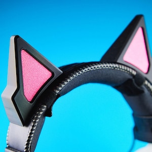 May include: Black and silver headset with cat ears. The headset features black ear cups with pink interior accents and silver trim. The headband is black and padded, with a silver zipper detail. The background is a solid blue.
