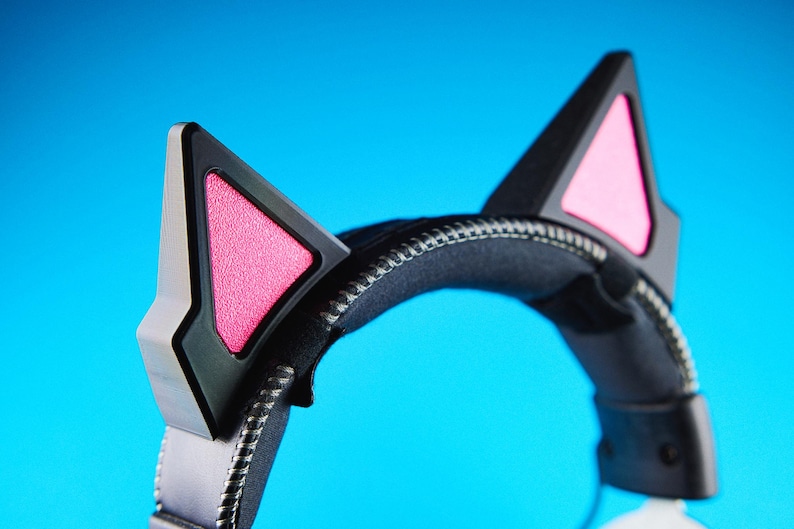 May include: Black and silver headset with cat ears. The headset features black ear cups with pink interior accents and silver trim. The headband is black and padded, with a silver zipper detail. The background is a solid blue.