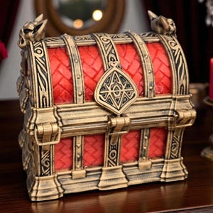 May include: A decorative treasure chest with a red interior and gold-colored accents. The chest features intricate detailing, including ornate carvings and a decorative clasp. The top of the chest has a series of arched supports. The chest is sitting on a wooden surface.
