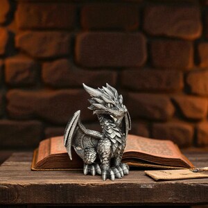 Puede incluir: Una figura de drag&oacute;n plateado con escamas y alas detalladas se encuentra sobre un libro abierto en una mesa de madera. El drag&oacute;n es el punto focal, con un tema de fantas&iacute;a medieval. El fondo presenta una pared de ladrillo y luz de velas.