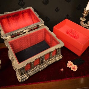May include: An open, ornate treasure chest with a red velvet interior sits on a red surface. A matching red box is next to it. The chest has a metallic finish with intricate detailing. Decorative candles and flowers are also present.