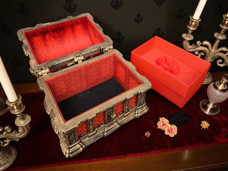 May include: An open, ornate treasure chest with a red velvet interior sits on a red surface. A matching red box is next to it. The chest has a metallic finish with intricate detailing. Decorative candles and flowers are also present.