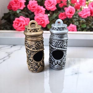 Puede incluir: Dos pequeños contenedores metálicos cilíndricos adornados con un diseño de huella de pata. Un contenedor es dorado y el otro es plateado. Ambos contenedores tienen una pequeña abertura en la parte superior.