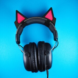 May include: Black over-ear headphones with cat ear attachments. The cat ears are black with pink inner ears. The headphones have a black headband and earcups, with a silver zipper detail on the headband. The background is a solid blue.