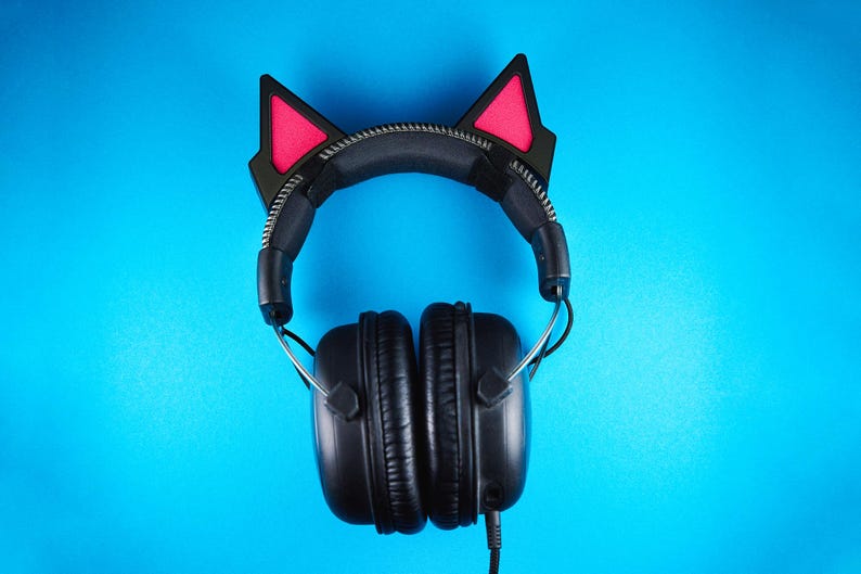 May include: Black over-ear headphones with cat ear attachments. The cat ears are black with pink inner ears. The headphones have a black headband and earcups, with a silver zipper detail on the headband. The background is a solid blue.