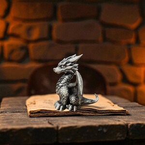 Puede incluir: Una detallada figura de drag&oacute;n plateado con intrincadas escamas y alas se encuentra sobre un libro antiguo y abierto. El drag&oacute;n est&aacute; posicionado sentado, con la cabeza girada hacia un lado. La escena se desarrolla sobre una superficie de madera r&uacute;stica contra un fondo de ladrillos.