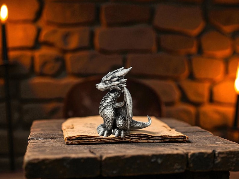 Puede incluir: Una detallada figura de drag&oacute;n plateado con intrincadas escamas y alas se encuentra sobre un libro antiguo y abierto. El drag&oacute;n est&aacute; posicionado sentado, con la cabeza girada hacia un lado. La escena se desarrolla sobre una superficie de madera r&uacute;stica contra un fondo de ladrillos.