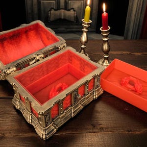 May include: An open, ornate chest with a red velvet interior and a matching small tray. The chest is silver and gold with intricate detailing. Two lit candlesticks with yellow and red candles are in the background.