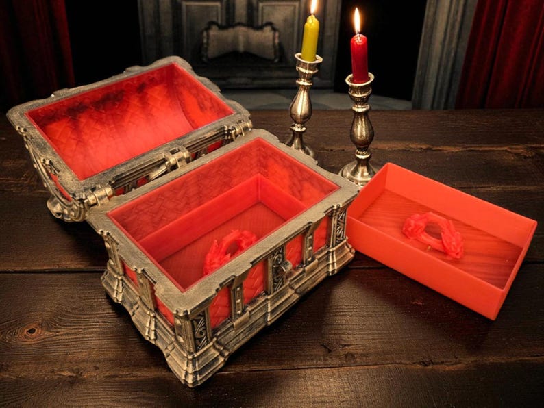 May include: An open, ornate chest with a red velvet interior and a matching small tray. The chest is silver and gold with intricate detailing. Two lit candlesticks with yellow and red candles are in the background.
