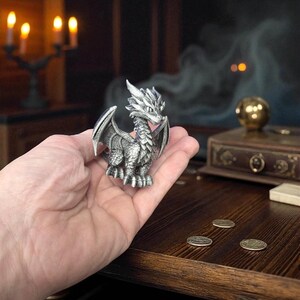 Puede incluir: Una detallada figura de drag&oacute;n plateado con alas extendidas, sostenida en una mano. El drag&oacute;n tiene escamas intrincadas y una cresta puntiaguda. El fondo incluye una mesa de madera, monedas y un entorno borroso y atmosf&eacute;rico con velas.